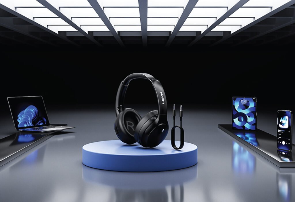 A pair of black LEVN headphones sit on a pedestal, surrounded by a laptop, tablet, and smartphones in a modern studio setting, showcasing the versatility of this Noise Cancelling Headphone.