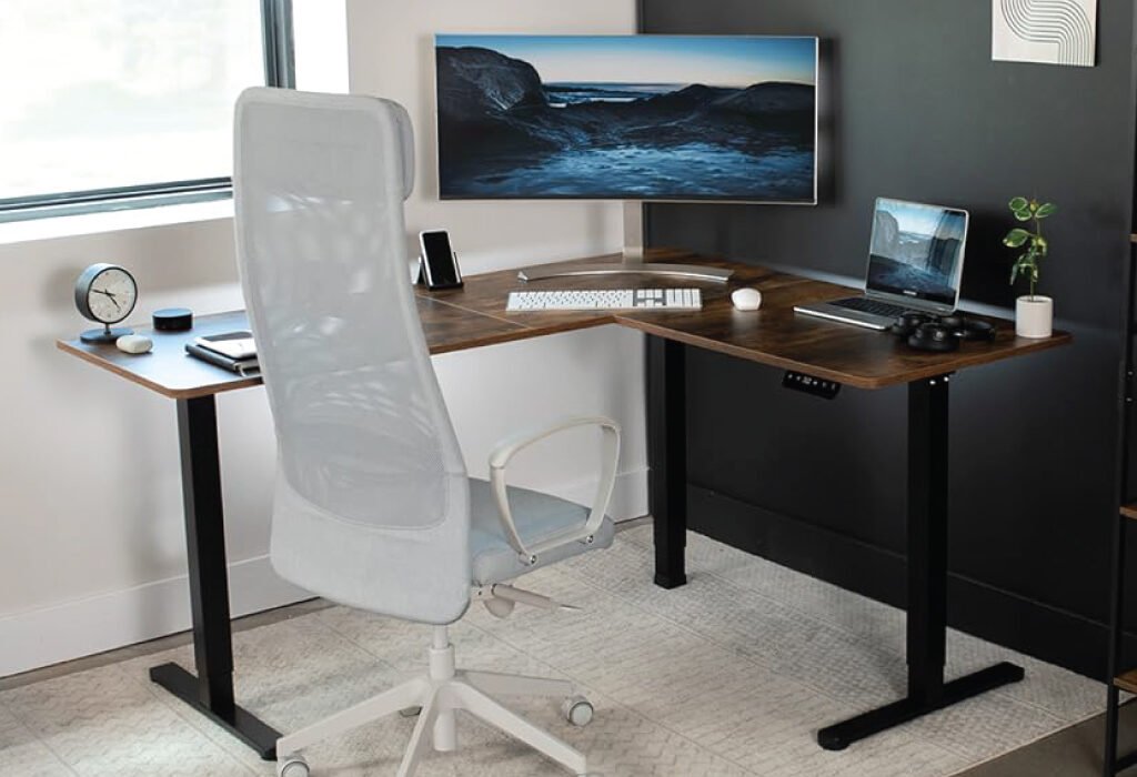 A corner office with a modern L-shaped Height Adjustable Desk in a home setting. The desk is equipped with a monitor, laptop, and a white ergonomic chair, showcasing a professional and comfortable workspace.